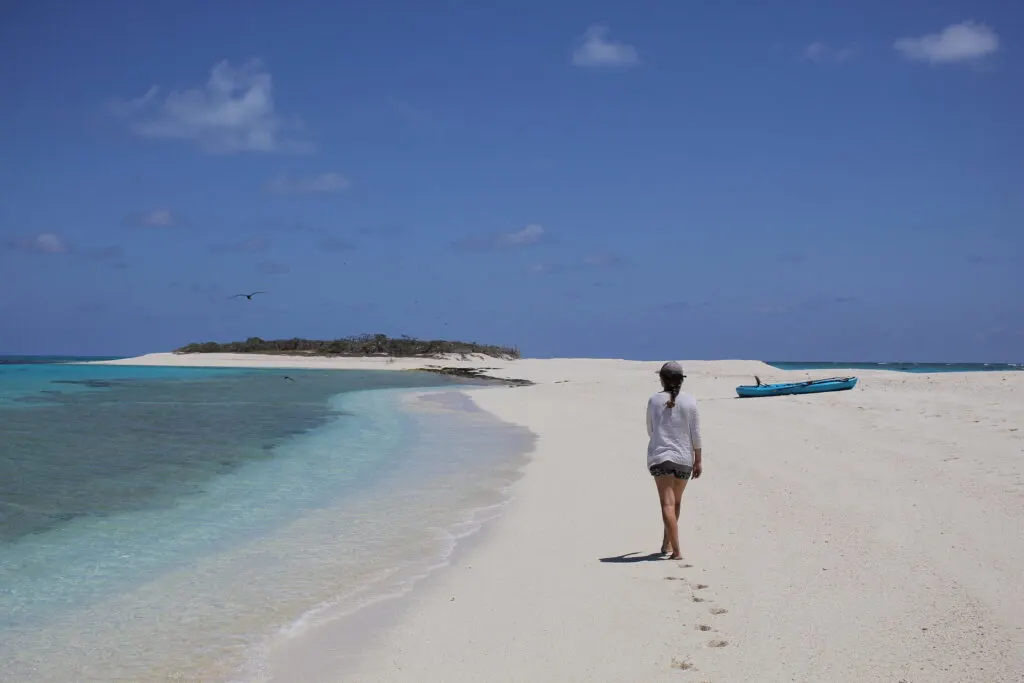 woman walking down white sand beach with blue kayak in background