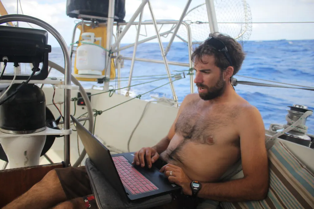 Man sits in sailboat cockpit reading laptop and looks surprised